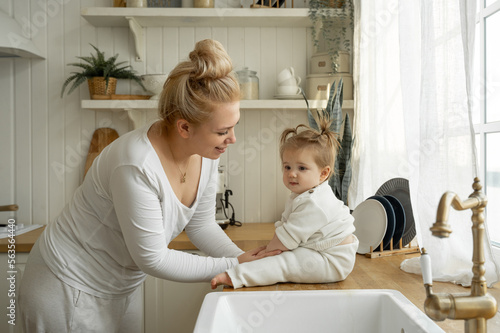 Mother Embrace And Kiss Baby Sitting On Table Kitchen. Domestic Life  Time Together  Maternity Leave Childhood. Mother s Day  Love Of Mom To Offspring Children.