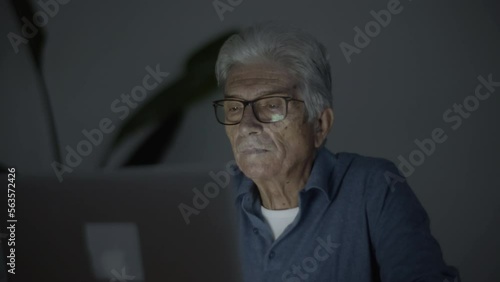 Tired senior man working on laptop in dark room. Medium shot of senior businessman typing on computer at night, looking at screen, solving business problems. Occupation, digital devices concept