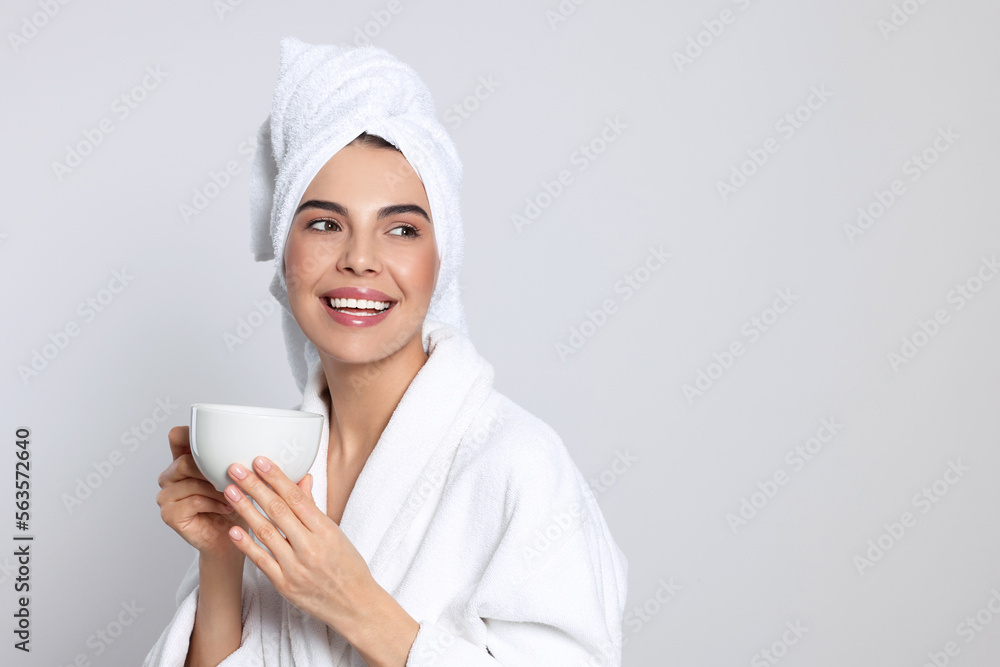 Young woman in bathrobe with towel and cup of drink on light grey background, space for text. Spa treatment Young woman in bathrobe with towel and cup of drink on light grey background, space for text. Spa treatment
