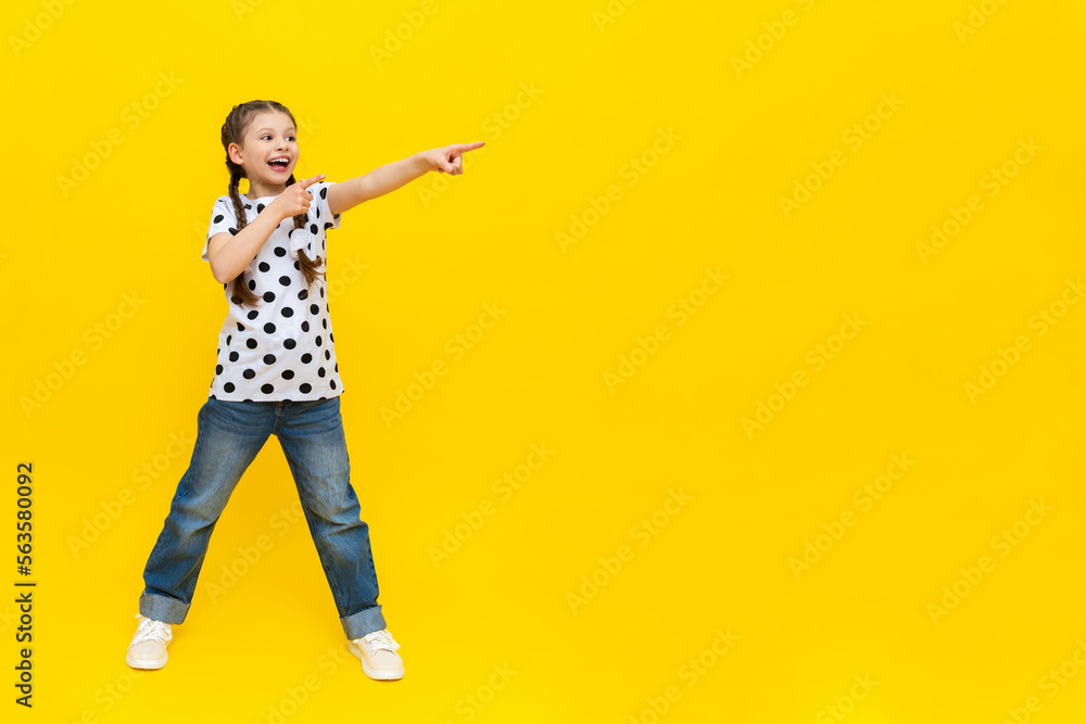A little girl with beautiful long pigtails in jeans on a yellow isolated background shows your advertisement. A little girl with beautiful long pigtails in jeans on a yellow isolated background shows your advertisement.