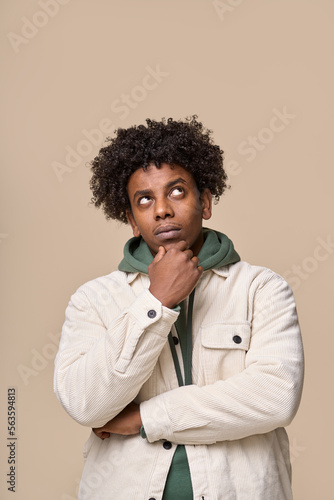 Young thoughtful doubtful African American guy student thinking of idea holding hand on chin, looking up choosing, making decision, feeling doubt having question isolated on beige background. Vertical