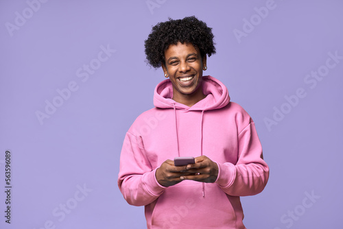 Happy cool curly African American teen guy model wearing pink hoodie holding cell phone using mobile digital apps on cellphone tech texting on smartphone isolated on light purple background.