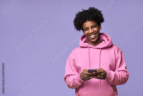 Slika na platnu Happy cool curly African American teenage guy teen boy model wearing pink hoodie holding cell phone using mobile digital apps on cellphone texting on smartphone isolated on light purple background