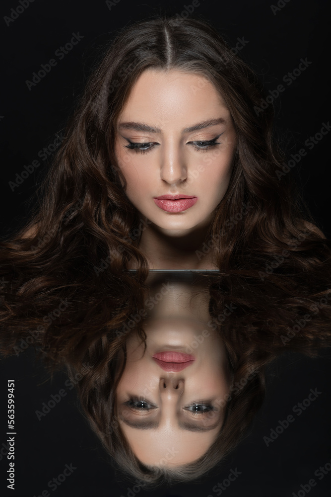 Make-up and fashion concept. Brunette woman with long wavy hair sensual studio portrait. Model looking to reflection of her face in mirror. Dark studio background