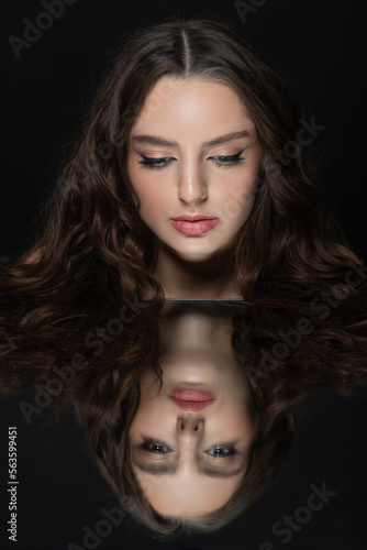 Make-up and fashion concept. Brunette woman with long wavy hair sensual studio portrait. Model looking to reflection of her face in mirror. Dark studio background