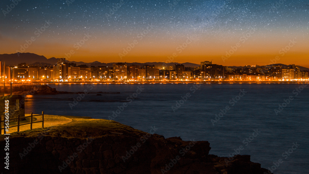 long exposition photo of a Starry night in the city of Gijon with the sea in the foreground and light contamination in the background
