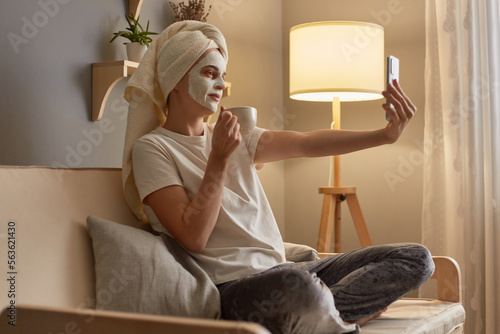 Image of beautiful woman in white T-shirt and pajama pants, wrapped in white towel sitting on sofa with cosmetic mask, doing cosmetology procedures, drinking coffee and making selfie on smart phone. photo