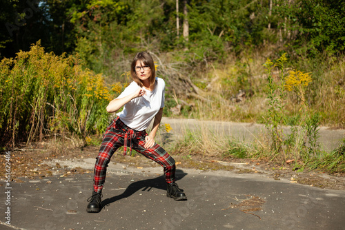 Outdoor-Portrait einer aktiven Frau, im weißem T-Shirt, karierte Hose mit hoher Taille trägt, auf einem Weg im Wald posiert.