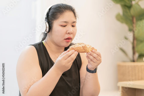 fat woman wearing headphones eating dessert after workout. chubby exerciser picking chocolate croissant wearing headset. overweight woman lost control to sweets gain unhealthy body listening to music