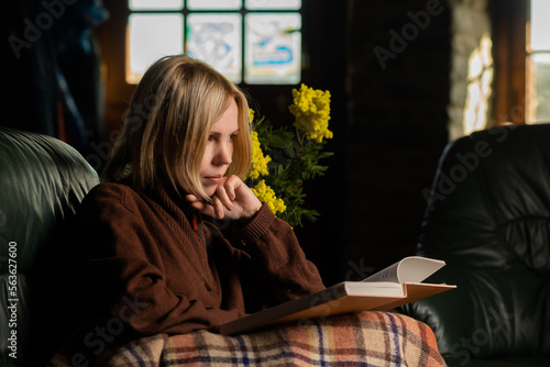 A middle-aged woman, forty years old, is reading a book on the couch. Woman in warm clothes covered with a blanket. Dark key photography.