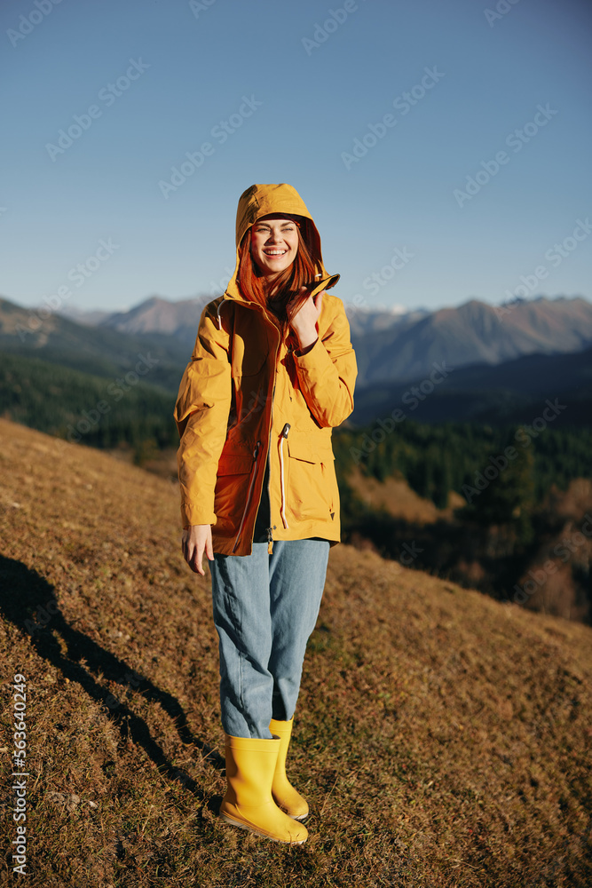 Woman in the autumn smile with teeth full-length walk on the hill and look at the mountains in a yellow raincoat and jeans happy sunset trip on a hike, freedom lifestyle 