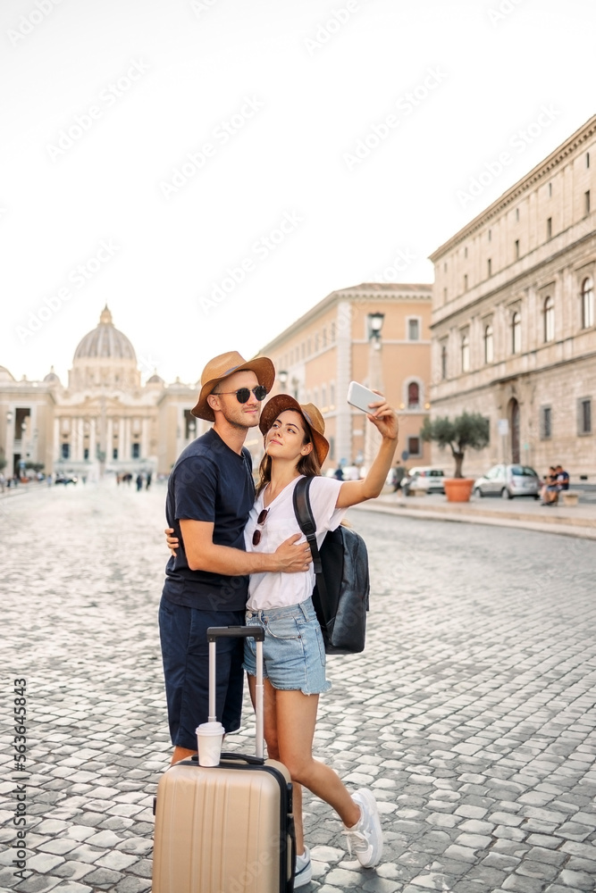 Happy young couple of travellers with hats and a suitcase hugging in the city and enjoying romance. Lovers smiling and have fun together. tourism. cheerful couple in casual clothes traveling the world