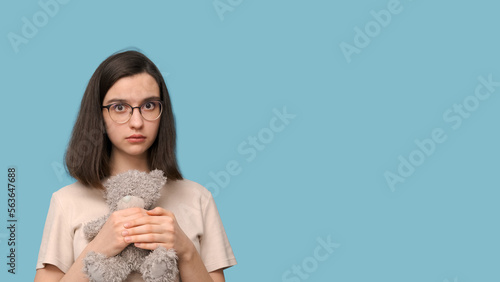 Portrait of a beautiful student girl with glasses, hugging a gray teddy bear toy on a blue background