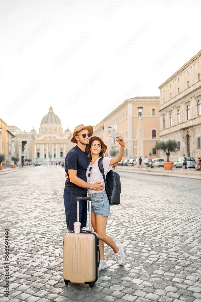 Happy young couple of travellers with hats and a suitcase hugging in the city and enjoying romance. Lovers smiling. A couple in love smiles and dreams
