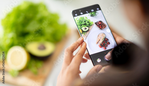 Young african american woman chef makes photo of fresh fish on smartphone screen at table with organic vegetables