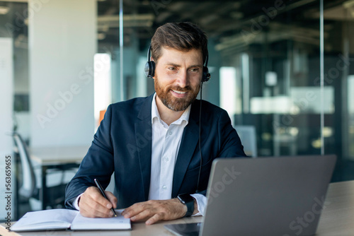 Happy middle aged male manager in headphones looking at laptop and making notes at workplace in office interior