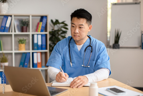Serious middle aged korean doctor in uniform with stethoscope looks at computer, makes notes at table