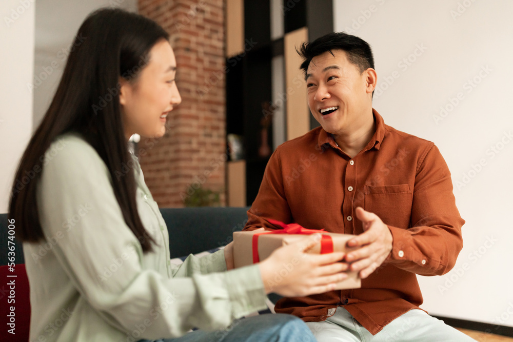Loving young asian wife giving present to her happy mature husband, sitting on couch at home Loving young asian wife giving present to her happy mature husband, sitting on couch at home