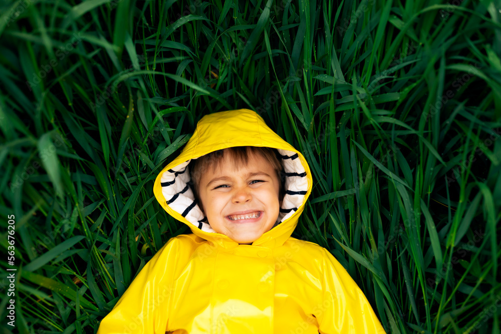 A little boy lies in the thick green grass in a yellow waterproof jacket and smiles.
