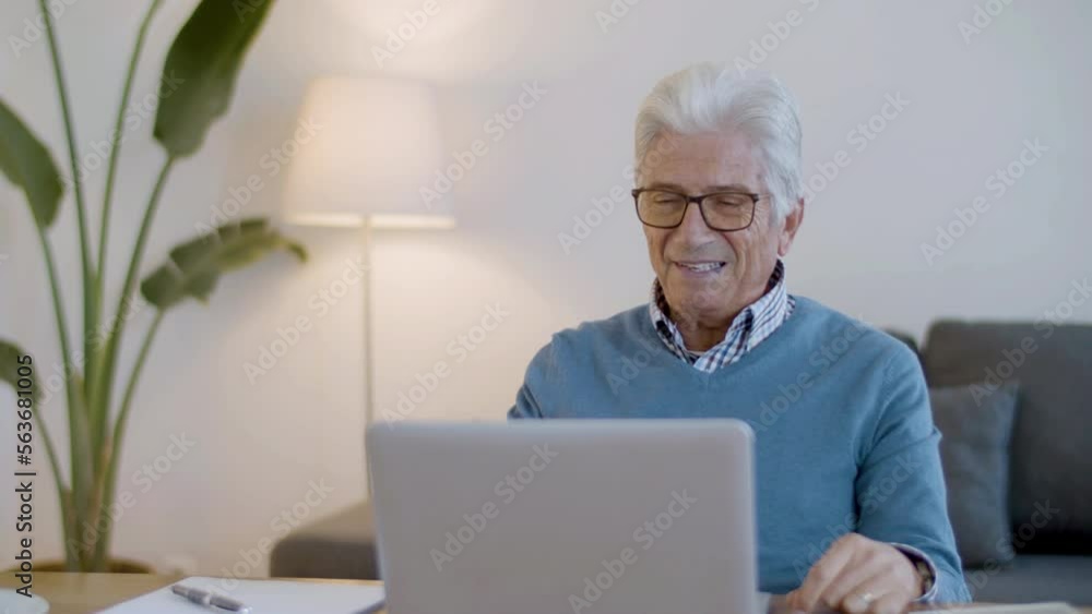 Happy senior man waving hand while having video call at home. Front view of cheerful mature man looking at laptop screen, sitting at table and talking with friends. Digital device, retirement concept Happy senior man waving hand while having video call at home. Front view of cheerful mature man looking at laptop screen, sitting at table and talking with friends. Digital device, retirement concept