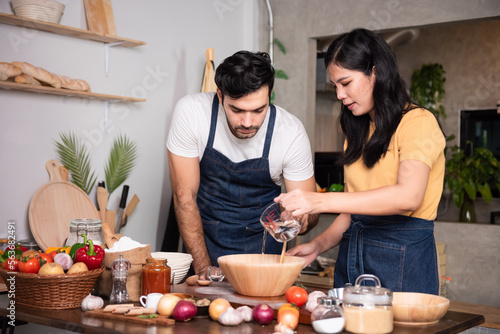 Happy family young couple chef preparing meal together in the kitchen. Caucasian man and asian women wearing aprons enjoy making pizza at home kitchen small business.