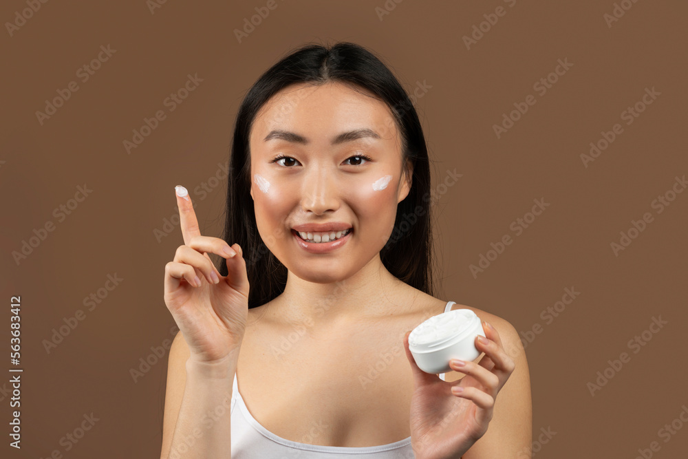 Happy young japanese lady holding jar of cream and smiling at camera, posing isolated on brown background, studio shot Happy young japanese lady holding jar of cream and smiling at camera, posing isolated on brown background, studio shot