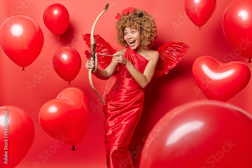 Happy positive female model pretends being cupid shoots with arrow at something wears red dress and wings comes on party for Valentines Day isolated over red background balloons flying in air
