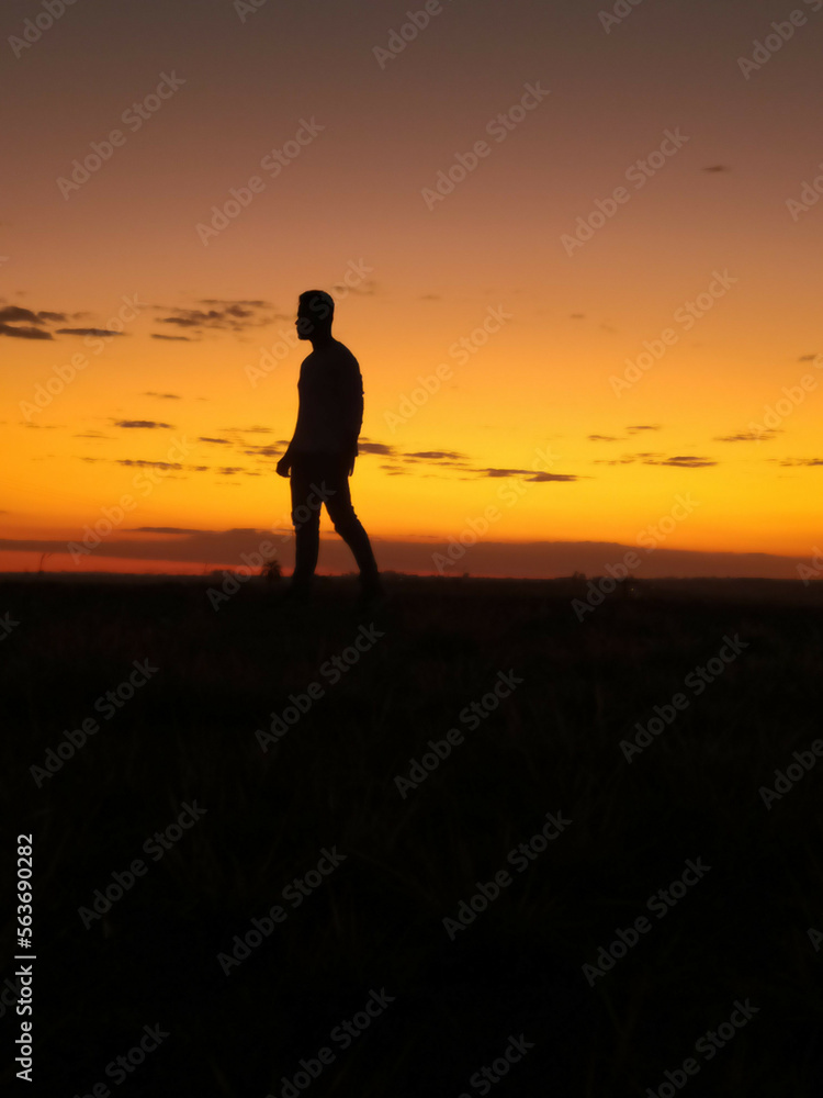 young man walking emotionless looking left silhouetted in sunset with orange sky in background