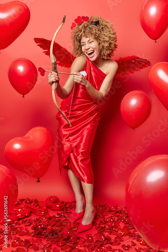 Vertical shot of cheerful woman with curly hair shoots with arrow and bow helps to find love for people wears long dress and wings behind back poses against red background inflated balloons.