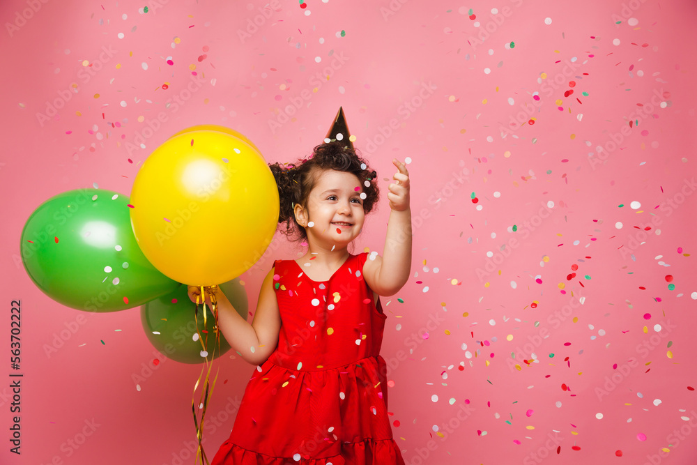 A joyful baby girl in a red dress celebrates her birthday and lets out colorful confetti on a pink background. a child holds balloons and catches confetti.