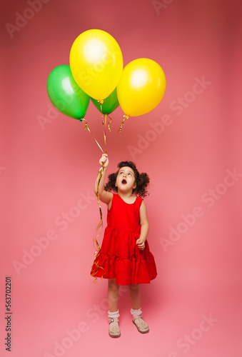 Portrait of a cheerful little girl isolated on a pink background, holding a bunch of colorful balloons, posing. © inna717