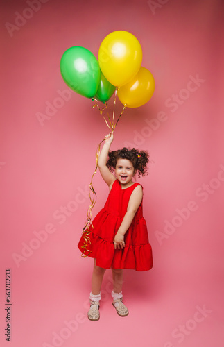 Portrait of a cheerful little girl isolated on a pink background, holding a bunch of colorful balloons, posing.