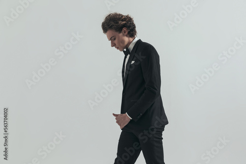 side view of blonde man with curly hair in black tuxedo walking and looking down