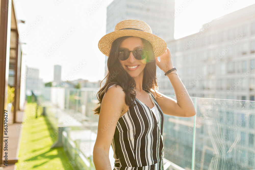 Funny charming woman with happy smile touching her hat and posing to camera in good mood. Pretty girl spend her weekend in the city in sunny day
