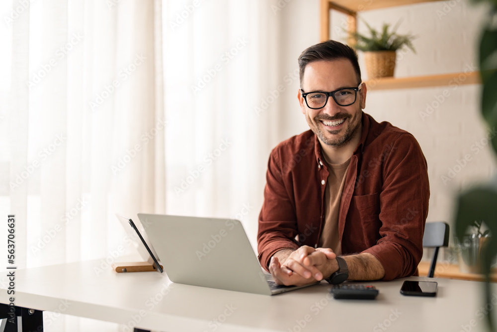 Smiling confident businessman looking at camera sitting at home office desk. Modern stylish corporate leader, successful CEO executive manager wearing glasses posing for business portrait. Smiling confident businessman looking at camera sitting at home office desk. Modern stylish corporate leader, successful CEO executive manager wearing glasses posing for business portrait.