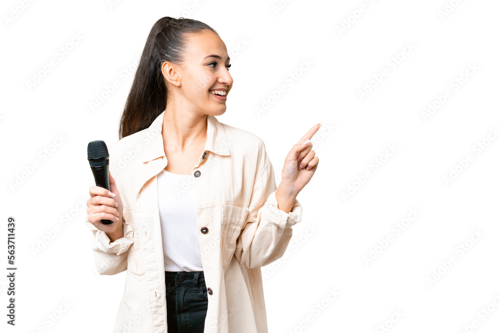 Young singer woman picking up a microphone over isolated chroma key background pointing finger to the side and presenting a product Young singer woman picking up a microphone over isolated chroma key background pointing finger to the side and presenting a product