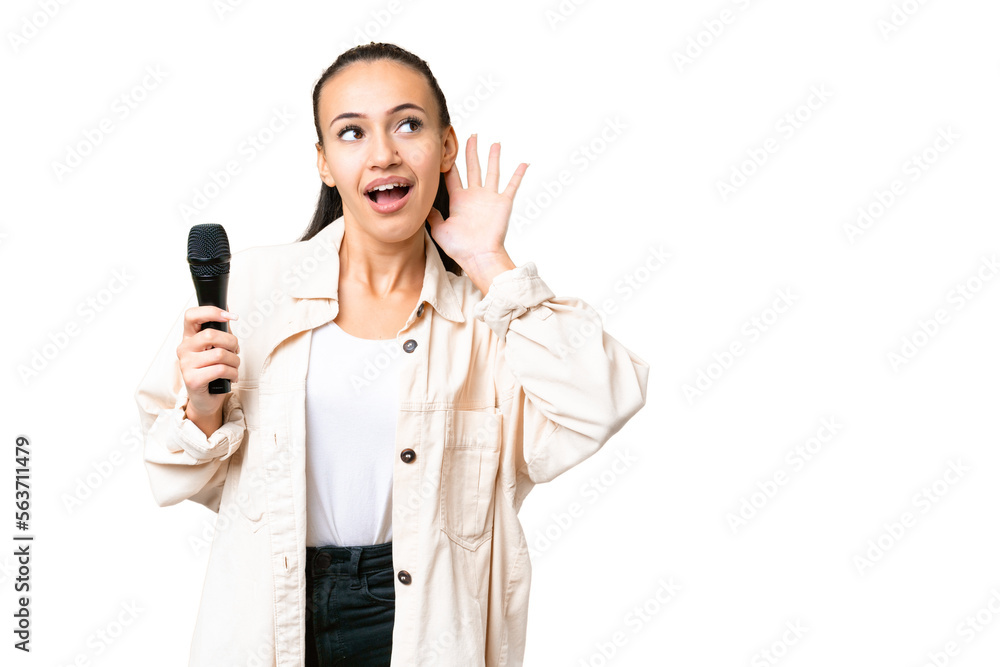 Young singer woman picking up a microphone over isolated chroma key background listening to something by putting hand on the ear Young singer woman picking up a microphone over isolated chroma key background listening to something by putting hand on the ear
