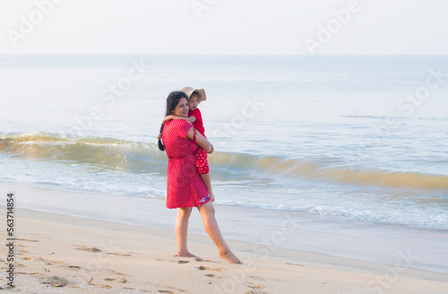 Attractive and Happy woman or lady in red dress with her daughter enjoys her tropical vacation on beach. Sand and blue water of Sea. Summer vacation and Holiday with Kid concept. Mother daughter Love.