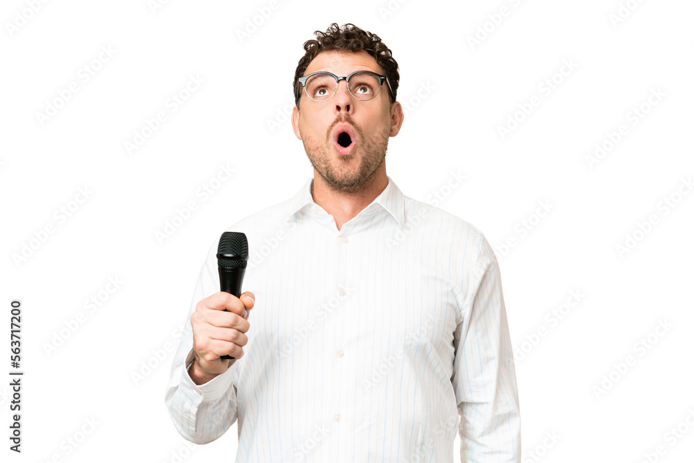Brazilian man picking up a microphone over isolated chroma key background looking up and with surprised expression Brazilian man picking up a microphone over isolated chroma key background looking up and with surprised expression