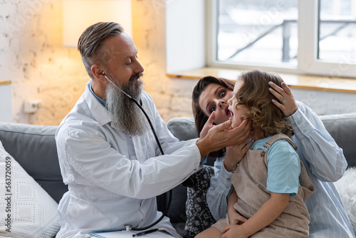 Fototapeta Naklejka Na Ścianę i Meble -  Kind male senior paediatrician doctor visiting his patient at home, examining little girl sitting on mother's lap, writing prescription. Concept of kid's health check