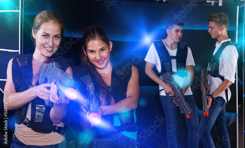 Two happy girls with laser guns in foreground and two guys in background in laser tag room