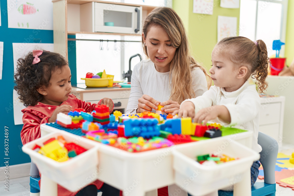 Obraz premium Teacher with girls playing with construction blocks sitting on table at kindergarten