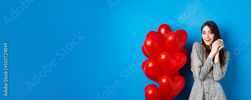 Valentines day. Lovely girlfriend ind ress smiling happy at camera, celebrating lovers holiday, standing near heart balloons, blue background photo