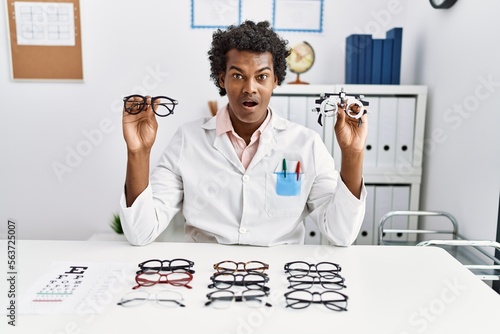 African optician man with curly hair holding optometry glasses and regular glasses afraid and shocked with surprise and amazed expression, fear and excited face.
