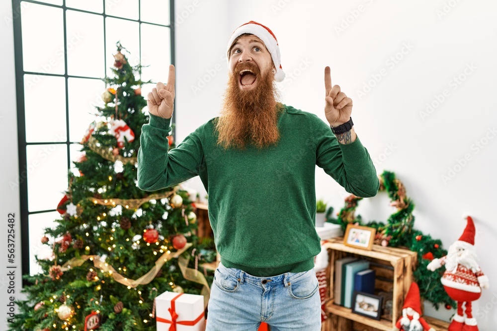 Redhead man with long beard wearing christmas hat by christmas tree smiling amazed and surprised and pointing up with fingers and raised arms.