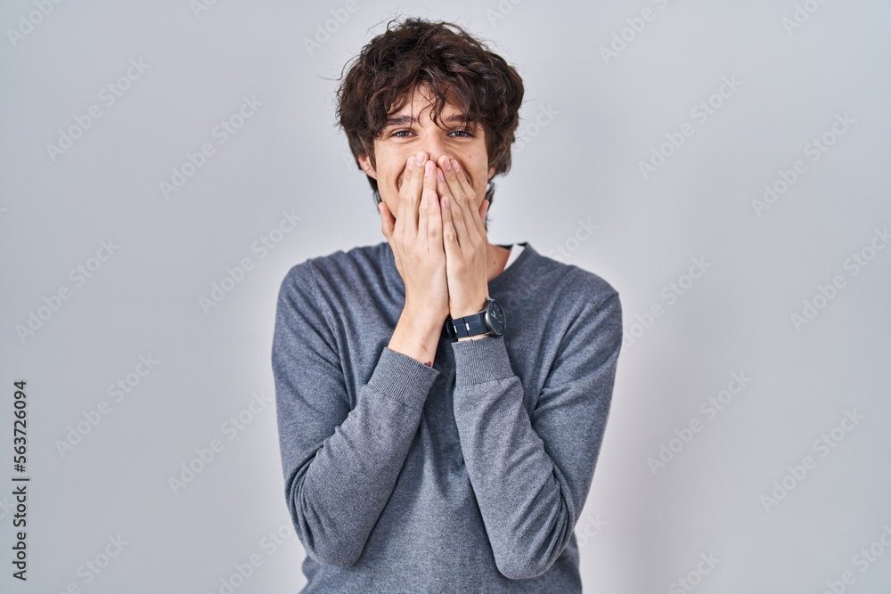 Young man standing over isolated background laughing and embarrassed giggle covering mouth with hands, gossip and scandal concept Young man standing over isolated background laughing and embarrassed giggle covering mouth with hands, gossip and scandal concept