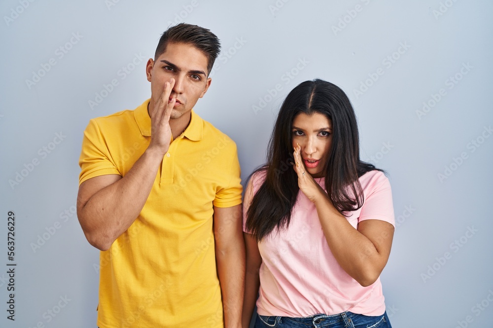 Naklejka premium Young couple standing over isolated background hand on mouth telling secret rumor, whispering malicious talk conversation