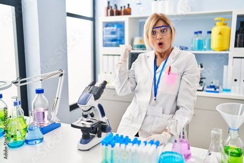 Middle age blonde woman working at scientist laboratory surprised pointing with hand finger to the side  open mouth amazed expression.