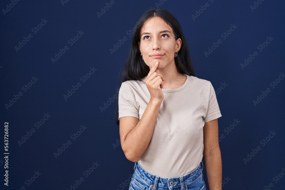 Young hispanic woman standing over blue background thinking concentrated about doubt with finger on chin and looking up wondering Young hispanic woman standing over blue background thinking concentrated about doubt with finger on chin and looking up wondering