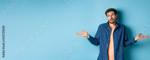 Indecisive young man with beard, look away and shrugging shoulders, know nothing, being clueless, standing on blue background photo